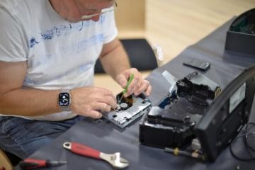 Ein Mann repariert ein Gerät. Zu sehen sind vor allem seine Hände, Werkzeug auf dem Tisch und eine offenes Gerät